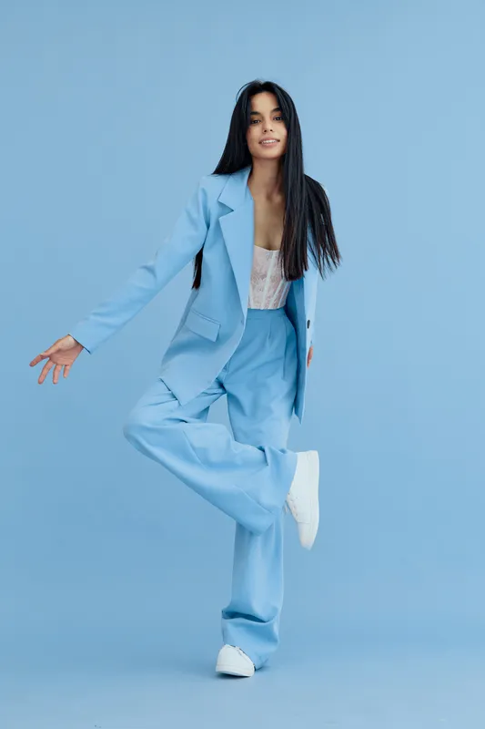 young woman posing in blue suit with white shoes on light blue background, casual fashion style, long dark hair, joyful expression, studio portrait