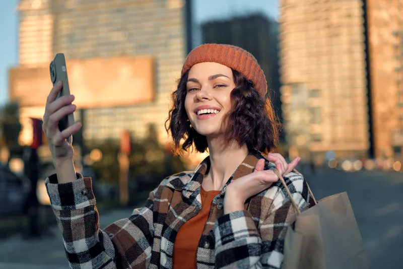 woman smiling selfie shopping urban beanie portrait: candid lifestyle scene of a young woman taking a phone selfie with shopping bags in city street at golden hour glow, authenticity and joy.