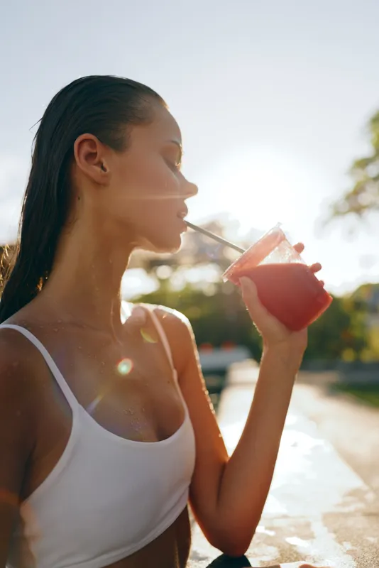 Young woman with wet hair drinks a fresh red smoothie through a straw outdoors in warm sunlight, enjoying a healthy lifestyle moment by the water.