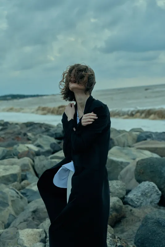 wind, sea, rocks, coast, woman, coat, fashion, moody, outdoors, windswept figure in a dark coat on rugged shoreline, dramatic seascape with stormy atmosphere, overcast sky, churning waves, distant