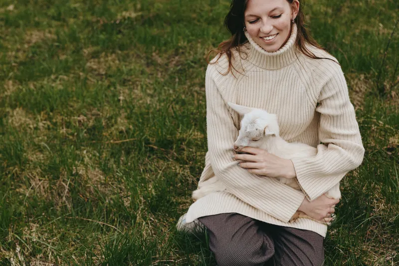 A young woman in a beige sweater gently holds a small white baby goat while sitting on grass in a natural outdoor setting, expressing warmth and tenderness in a casual lifestyle moment.