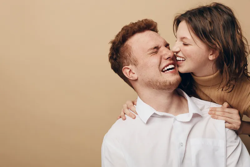 Happy young couple laughing together in casual clothes on beige background. Close emotional moment shows love, joy, and connection between man and woman in warm light.