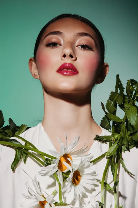 Close-up portrait of confident young woman with bold red lipstick and flushed cheeks, adorned with daisies on white outfit. Concept of beauty, fashion, creativity, and freshness.