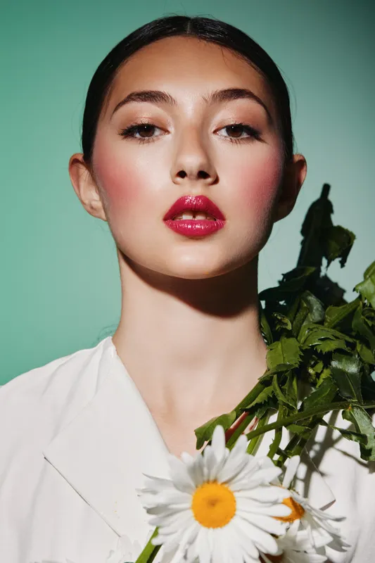 Close-up of a young woman with glowing skin and bold makeup holding daisies. Green background contrasts with her vivid red lips and soft natural flower. Beauty and fashion concept.
