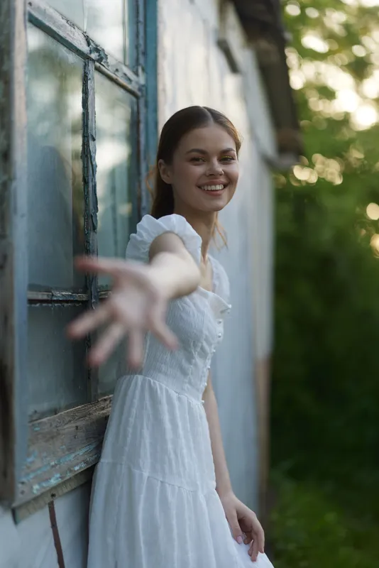 Young woman in a white dress reaching out, smiling warmly against a blurred natural background in soft daylight, evoking warmth and connection during springtime