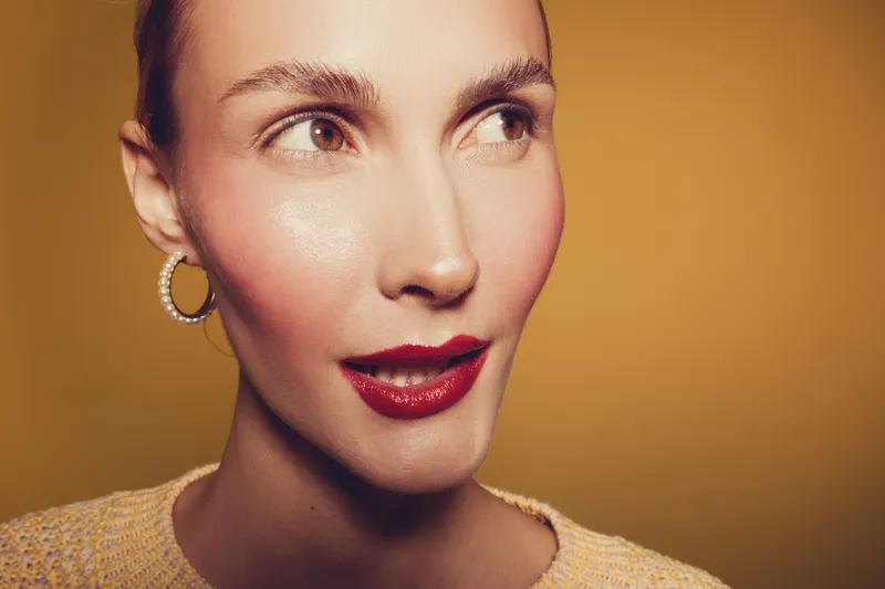 Close-up portrait of a woman with red lipstick and hoop earrings looking sideways on a warm brown background. Concept of beauty, makeup, fashion, and confident expression.