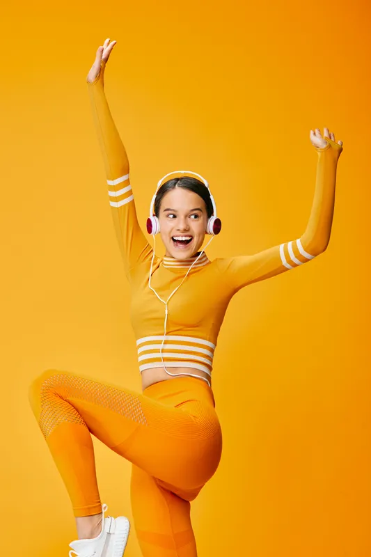 Dynamic woman in orange workout outfit jumps with raised arms, wearing headphones, against a vibrant backdrop, capturing fitness energy, sport performance, and joyful dance.