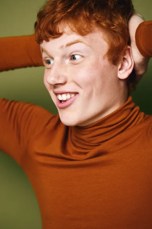 Young man with red hair and freckles wearing orange turtleneck sweater looking excited and happy with hands behind head on green background. Casual expression and natural smile.