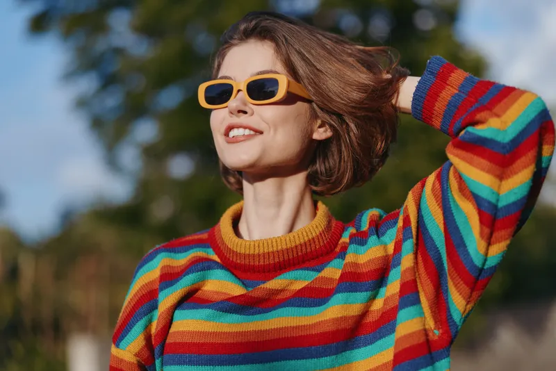 Woman, smiling, rainbow sweater, yellow sunglasses, short hair, outdoor, sunny, casual: Young 20s female model in colorful striped sweater, happy and confident, looking up with hand in hair in park.