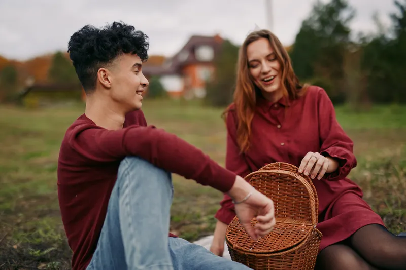 A candid young couple sharing a relaxed outdoor picnic with a wicker basket, smiling and engaging in a genuine, authentic moment that highlights realism and natural connection.