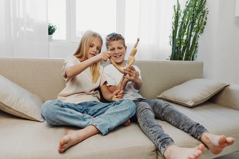 Two happy children sitting on a beige sofa playing with a wooden mannequin doll in a bright living room with natural light and plants. Concept for childhood fun and creativity.