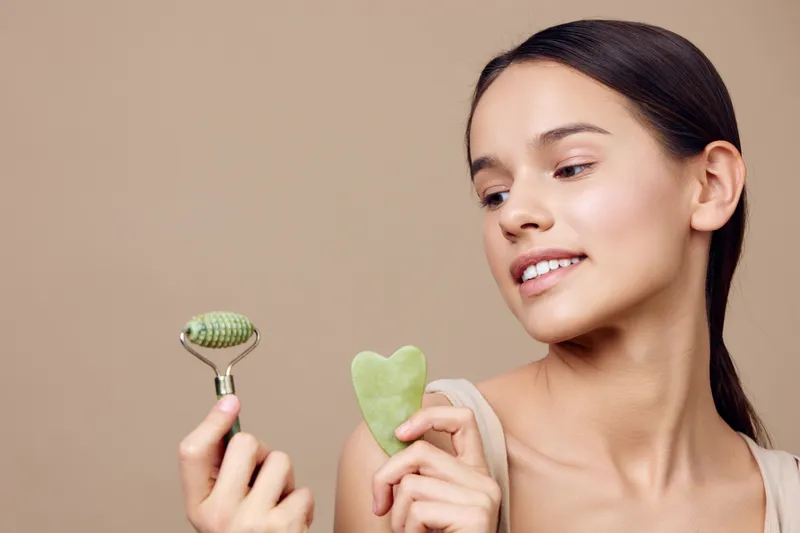 Young woman holding jade roller and gua sha tool for facial massage, promoting self care and beauty routines in a calm, neutral background