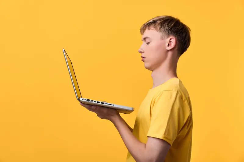 Boy in a yellow shirt stands in profile, holding a laptop open, focused on the screen for digital learning, education and online study, bright studio backdrop, modern tech education mood, ready for