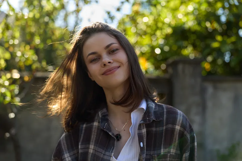 Cheerful woman outdoors, smiling with relaxed posture, sunlit moment, casual plaid shirt, natural colors, soft breeze, lush greenery backdrop