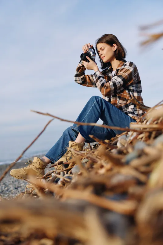Photographer in plaid shirt captures outdoor scenery, sitting on a rocky hillside among autumn leaves with a camera, focusing on composition and natural light for creative nature photography.