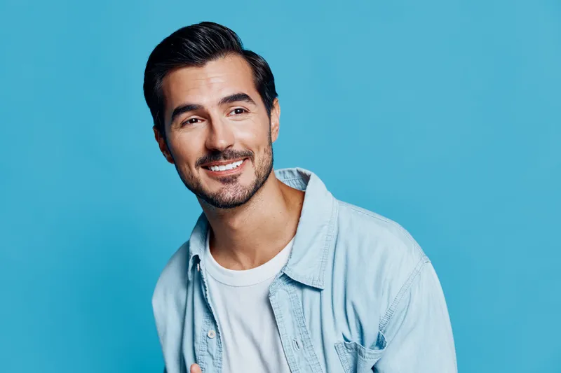 Happy smiling man in light denim shirt looking at the camera with a bright smile against a solid turquoise background. Cheerful lifestyle concept.