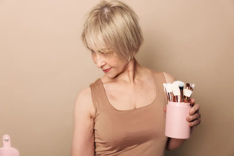 Mature woman with short blonde hair wearing a beige tank top holds a pink container filled with makeup brushes. Neutral background emphasizes beauty tools and skincare routine concept.