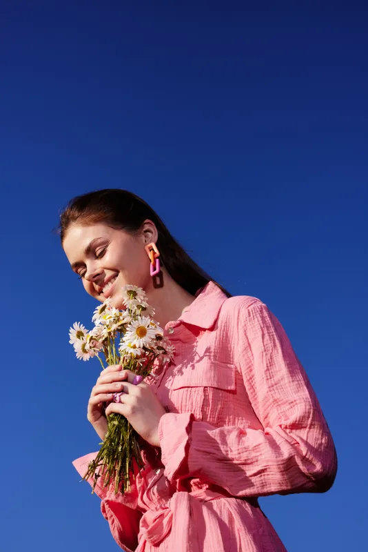 Young woman in a pink dress holding daisies against a blue sky, radiating joy and confidence during a bright, sunny day