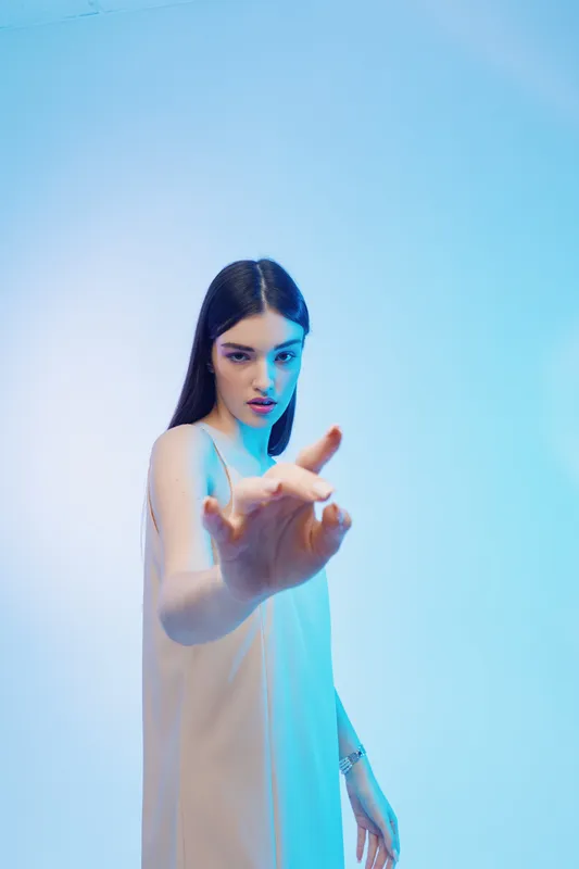 Young woman with long dark hair in a light dress reaches forward with hand. Studio shot on blue background. Concept of beauty fashion and elegance.