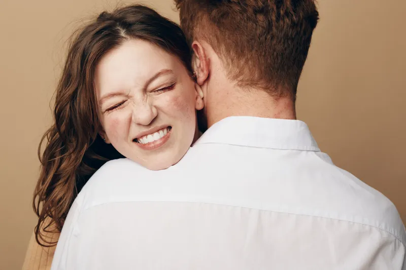 Young woman with long hair hugs man wearing white shirt, eyes closed and smiling. Intimate moment of affection and love on beige background, concept of relationship and happiness.