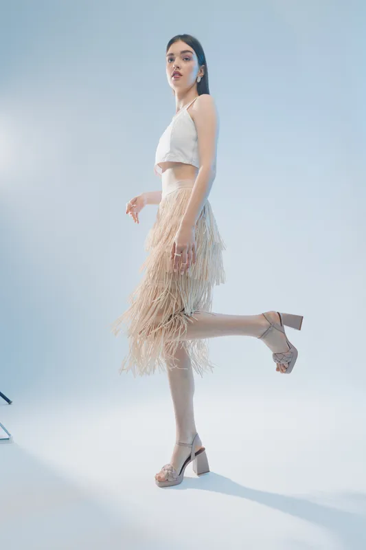 Young woman in a stylish beige fringe skirt and white crop top posing on one leg against a light background. Concept for woman beauty fashion and modern style.
