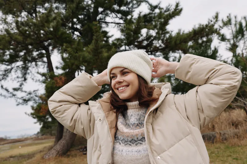 Woman in a warm coat and beanie outdoors, smiling as she adjusts her hood in a chilly rural setting