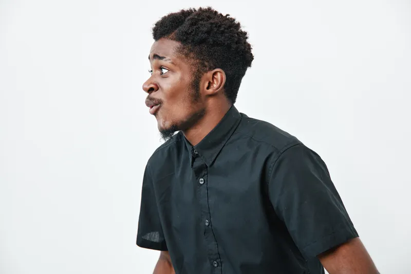 Young black man with dark skin and curly hair wearing black shirt looking surprised or amazed on white background. Side profile portrait of African ethnicity male with expressive face.