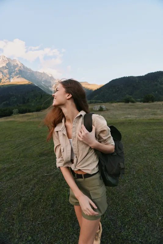 Young woman with a backpack walks through a sunlit meadow beside distant mountains, smiling softly while admiring the expansive landscape