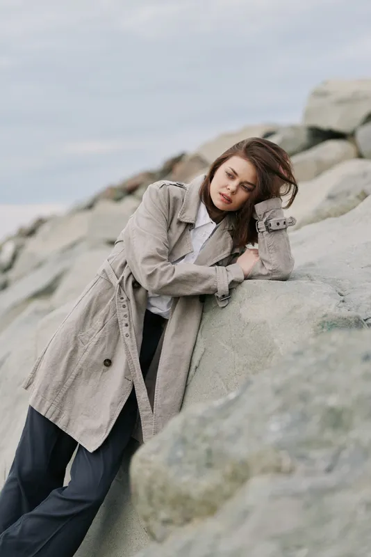 Young woman in knitted coat leans on rocks outdoors, eyes closed, enjoying peaceful moment. Calm nature setting with muted colors and soft natural light.