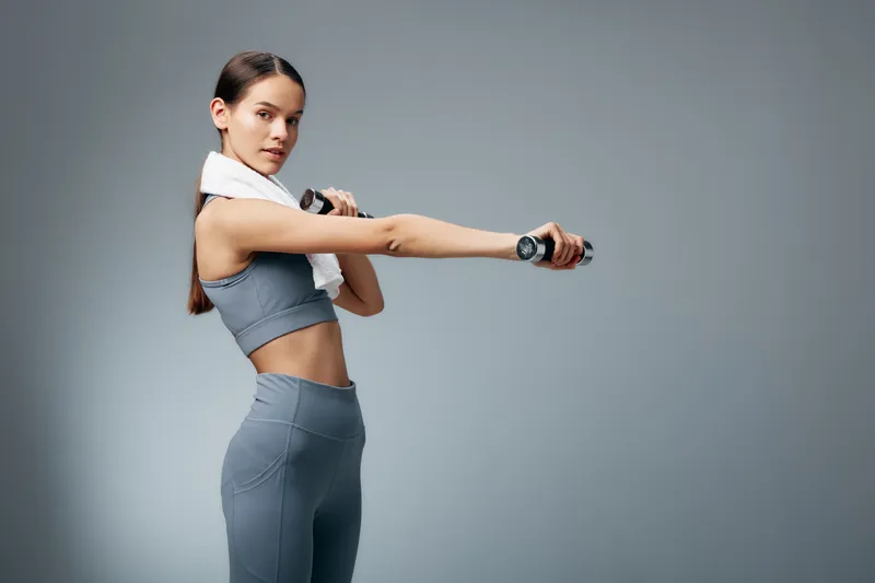 Woman fitness athlete performing a stretching exercise with dumbbells, towel over her shoulder, wearing gray sportswear in a minimal studio for sport training and healthy lifestyle.