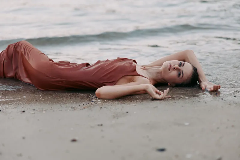 A woman lies on damp sand by the quiet sea, wearing a flowing earthy dress. Soft twilight light caresses her serene pose, while gentle waves frame a tranquil, intimate scene.
