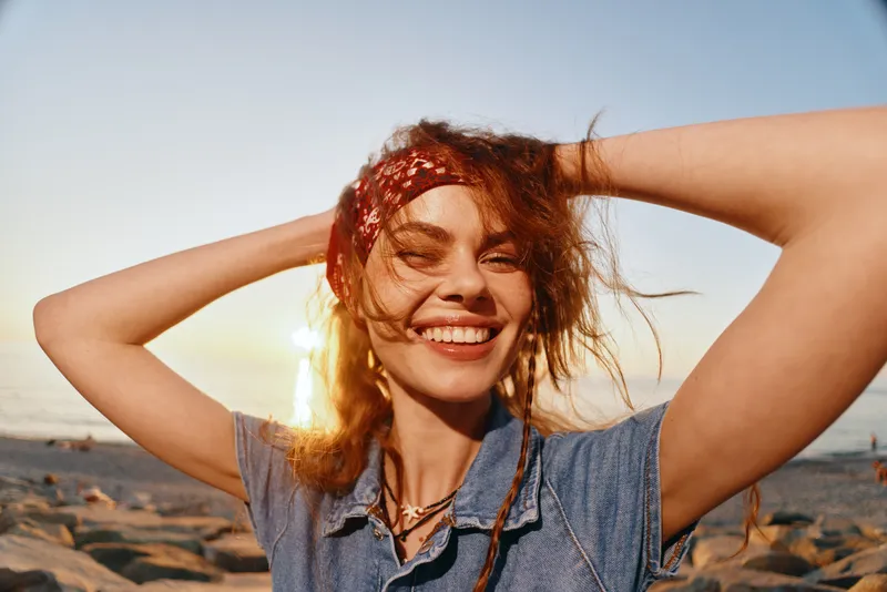 smiling woman enjoying sunset at the beach with carefree expression and red bandana hair accessory, warm natural light and casual blue denim shirt