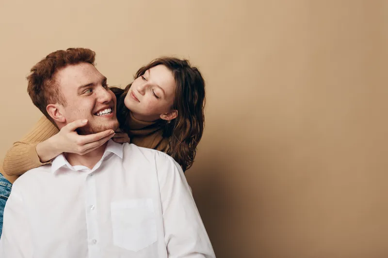Happy young couple embraces and smiles, woman gently holding man's face. Warm beige background creates cozy atmosphere. Concept of love, affection, and romantic relationship.