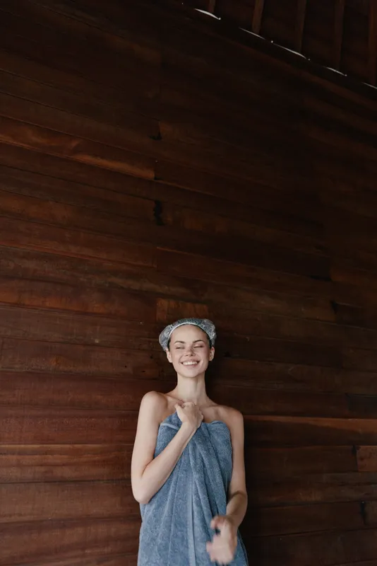 smiling woman wrapped in towel with headband stands against wooden wall indoors, expressing relaxation and calmness in a cozy spa environment.