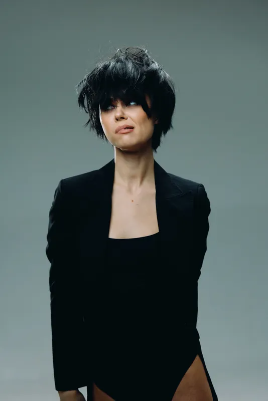 woman with short black hair in black outfit posing confidently against gray background