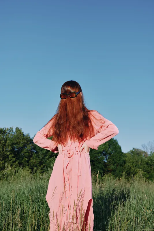 Woman in a flowing pink dress standing confidently in a green field with long red hair covering her face, capturing a sense of empowerment and mystery