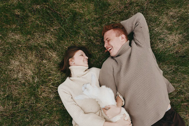 Cheerful couple outdoors, lying on grass, sharing a warm moment with a playful goat kid nearby, cozy sweaters, natural light, relaxed mood, candid interaction, tranquil colors, lifestyle scene