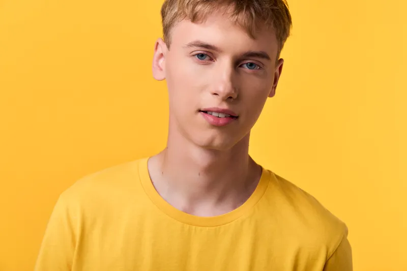 Young man in a yellow shirt stands against a vibrant yellow background, showcasing casual fashion and a friendly expression in a headshot style for lifestyle or fashion projects