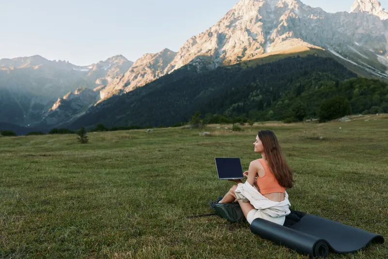 A woman sits on a rolled mat in a grassy meadow, using a laptop with a dramatic mountain backdrop. She smiles slightly, focused on work, creating a calm outdoor office mood.