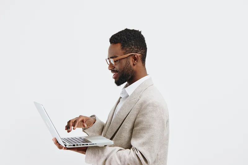 Professional African-American man wearing glasses and beige blazer holding a laptop, smiling confidently against plain white background. Studio portrait of a businessman or professional working. Peopl