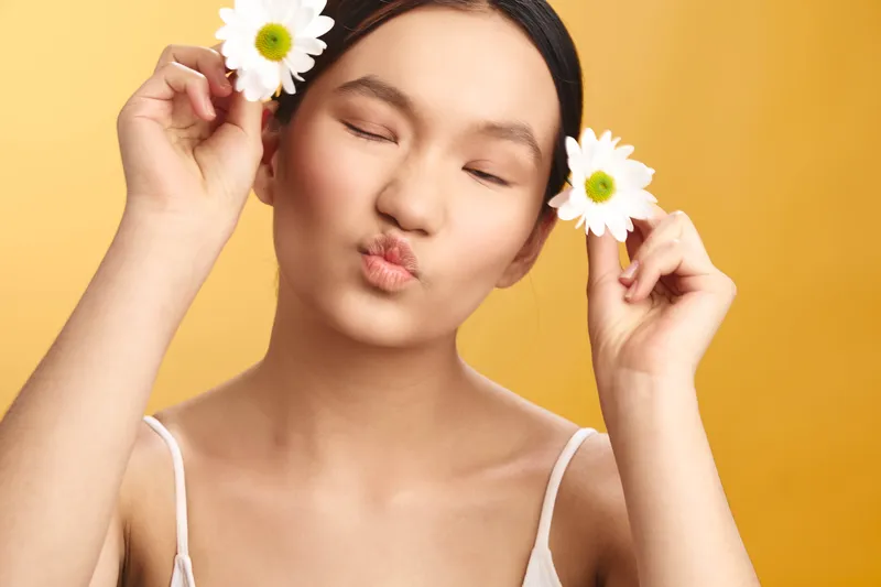 Asian woman exudes joy and calm as she poses with fresh daisies against a warm studio backdrop, her eyes closed and lips puckered in a gentle, playful kiss.