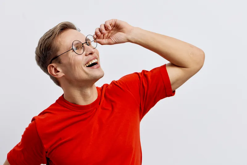Smiling young woman with short hair and glasses wearing a bright red t-shirt, laughing joyfully while adjusting her glasses, isolated on plain white background, studio portrait, cheerful, casual lifes