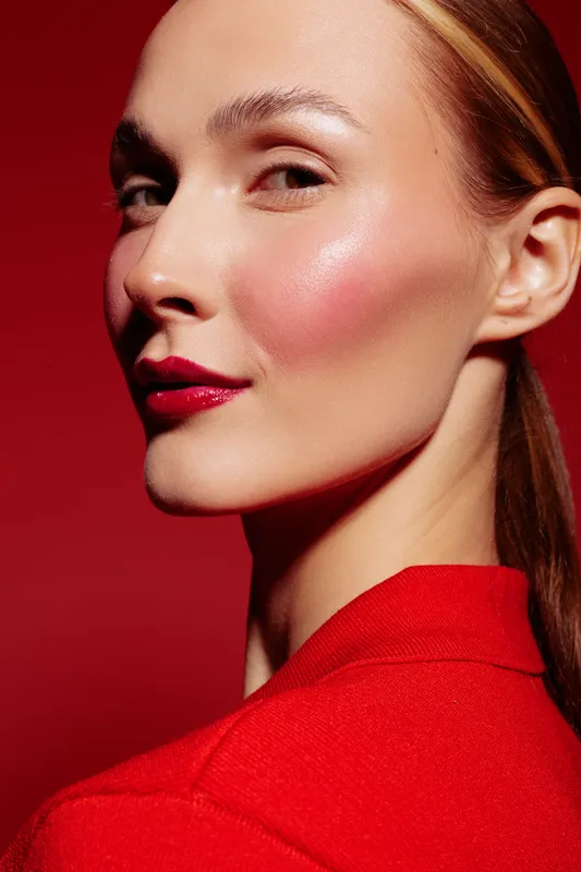 Close-up portrait of confident woman with glossy red lips and glowing skin wearing red outfit on red background. Concept of beauty, makeup, fashion, and elegance in studio lighting.