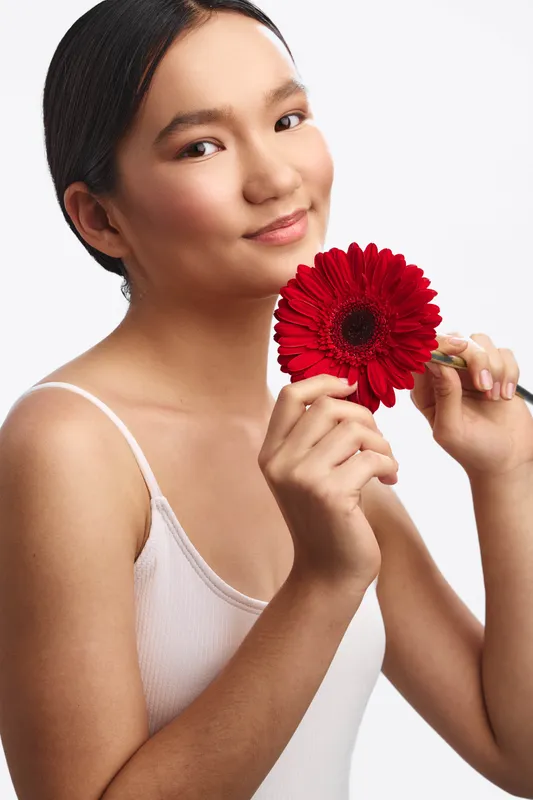 asian woman gently holds a bright red flower near her face in a simple studio setting, exuding calm elegance and soft confidence.