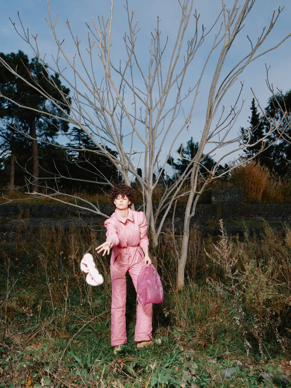Woman in a pink jumpsuit standing in a field beside a bare tree, holding a pink bag and a shoe, daytime outdoor scene, rural landscape, fashion pose in natural surroundings.