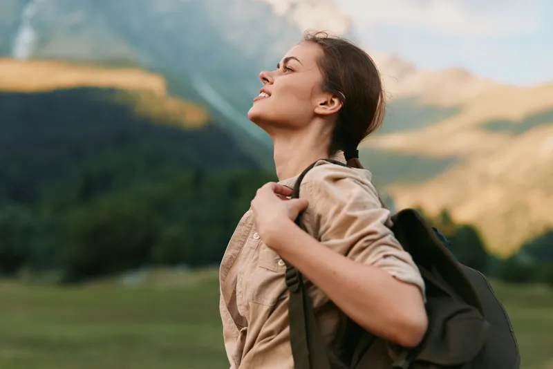 Young woman with backpack walks through a wide green valley as rugged mountains loom in the distance, embodying adventure, exploration and freedom.