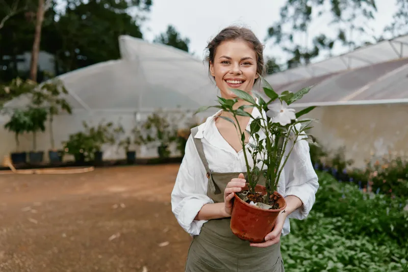 Young woman smiling while holding a potted plant in a greenhouse, showcasing a passion for gardening and a love for nature in a vibrant, green environment