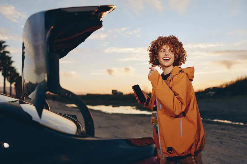 Woman lifestyle outdoors near car at sunset, curly haired woman laughing and smiling, orange jacket, holding smartphone, warm golden light, relaxed travel moment, beach background