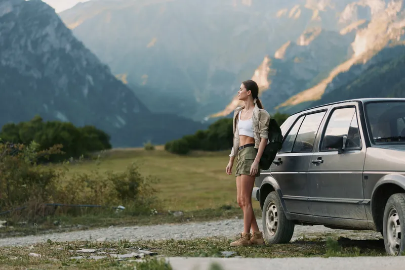 Smiling woman in casual outfit standing beside a car in a mountain landscape. Outdoor lifestyle, nature, travel vibe with warm light and wide scenery. Soft lighting enhances mood of freedom,