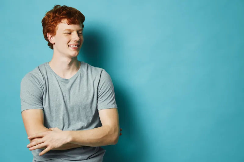 Young man with red hair and casual grey t shirt smiling with eyes closed standing against blue background for happiness and relaxation concept portrait.